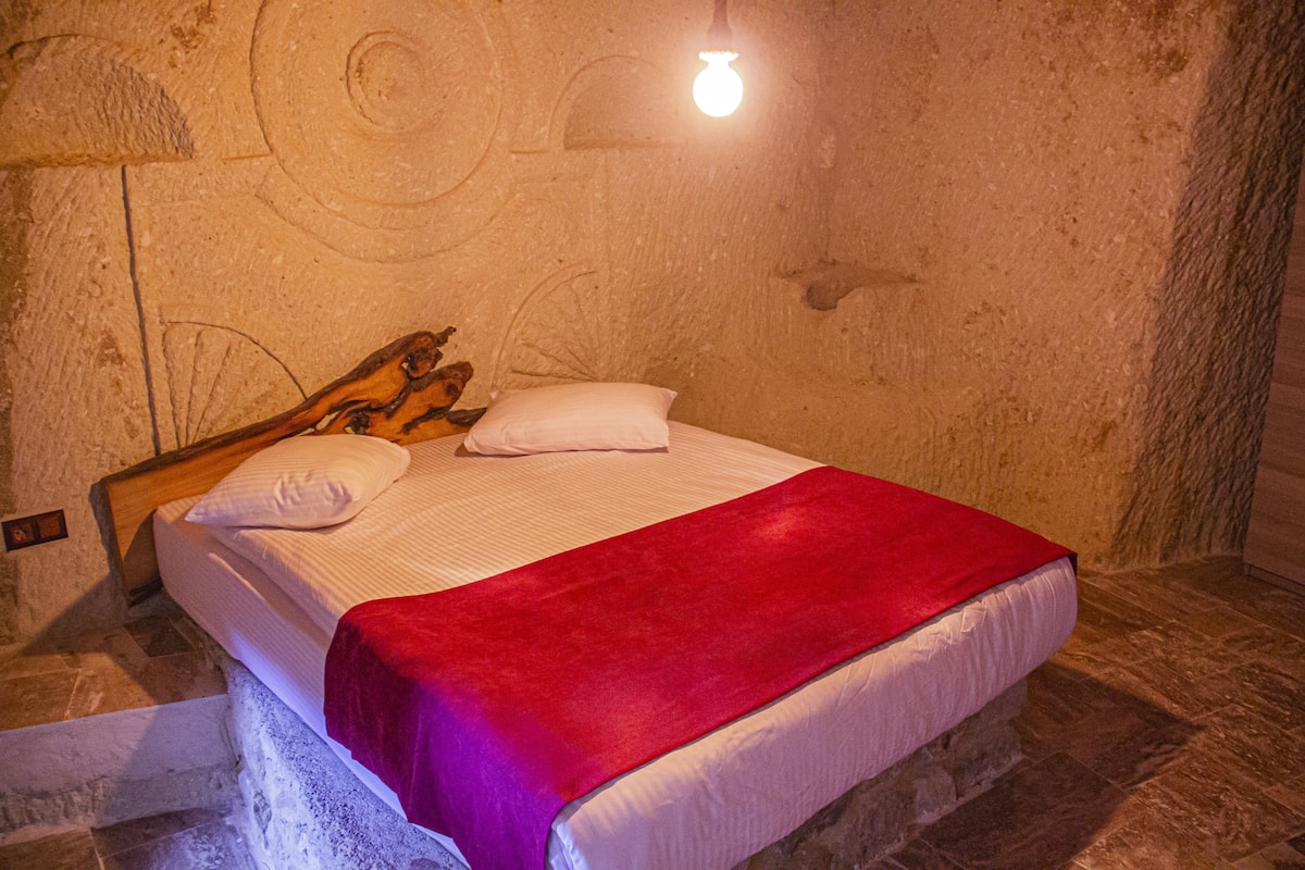 Una cama con sábanas blancas y una colcha roja, un cabecero de madera y una pared de piedra con una lámpara.