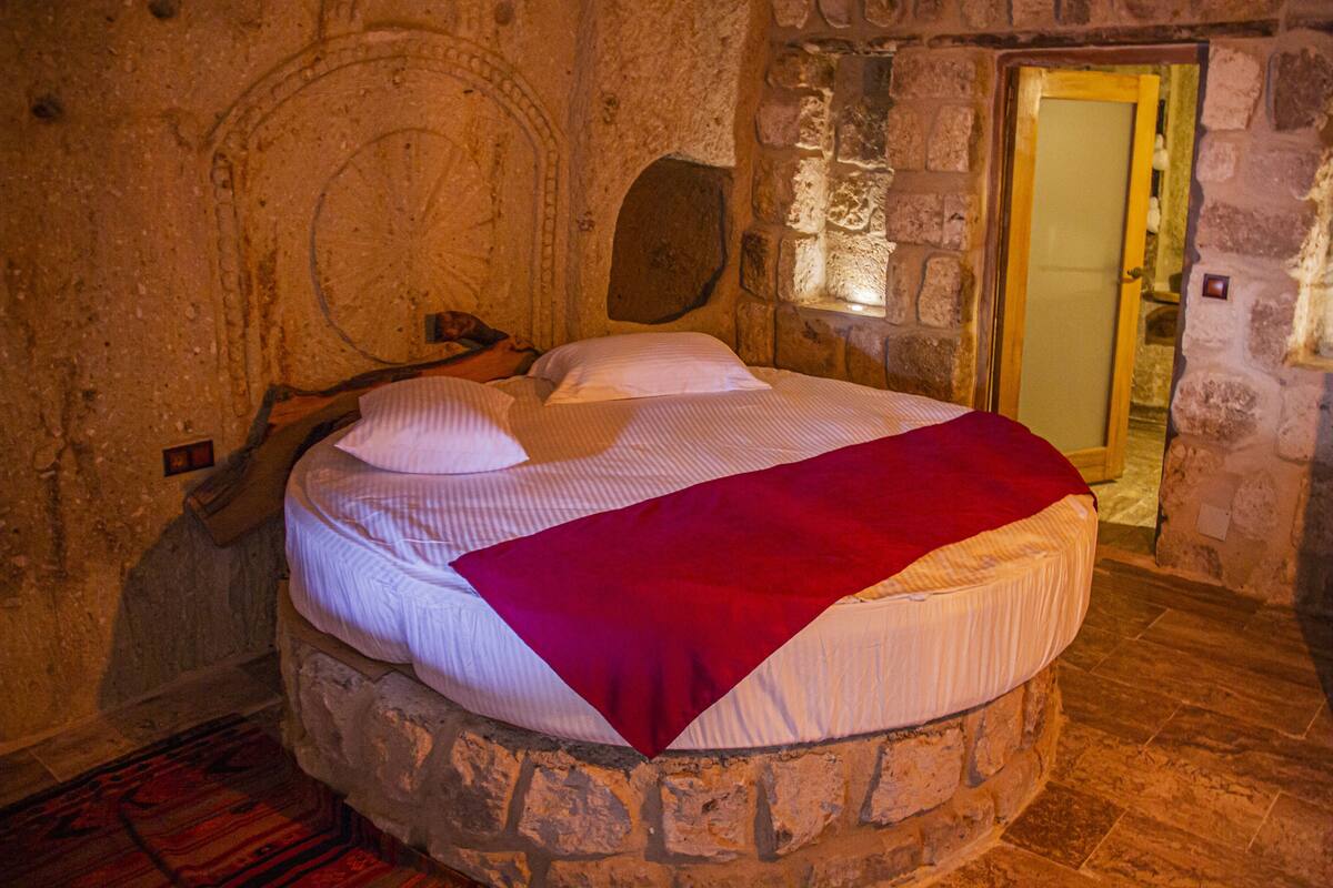 Una habitación de paredes de piedra con una cama ubicada en un nicho circular de piedra, una puerta de madera y un banco de piedra adosado a la pared.