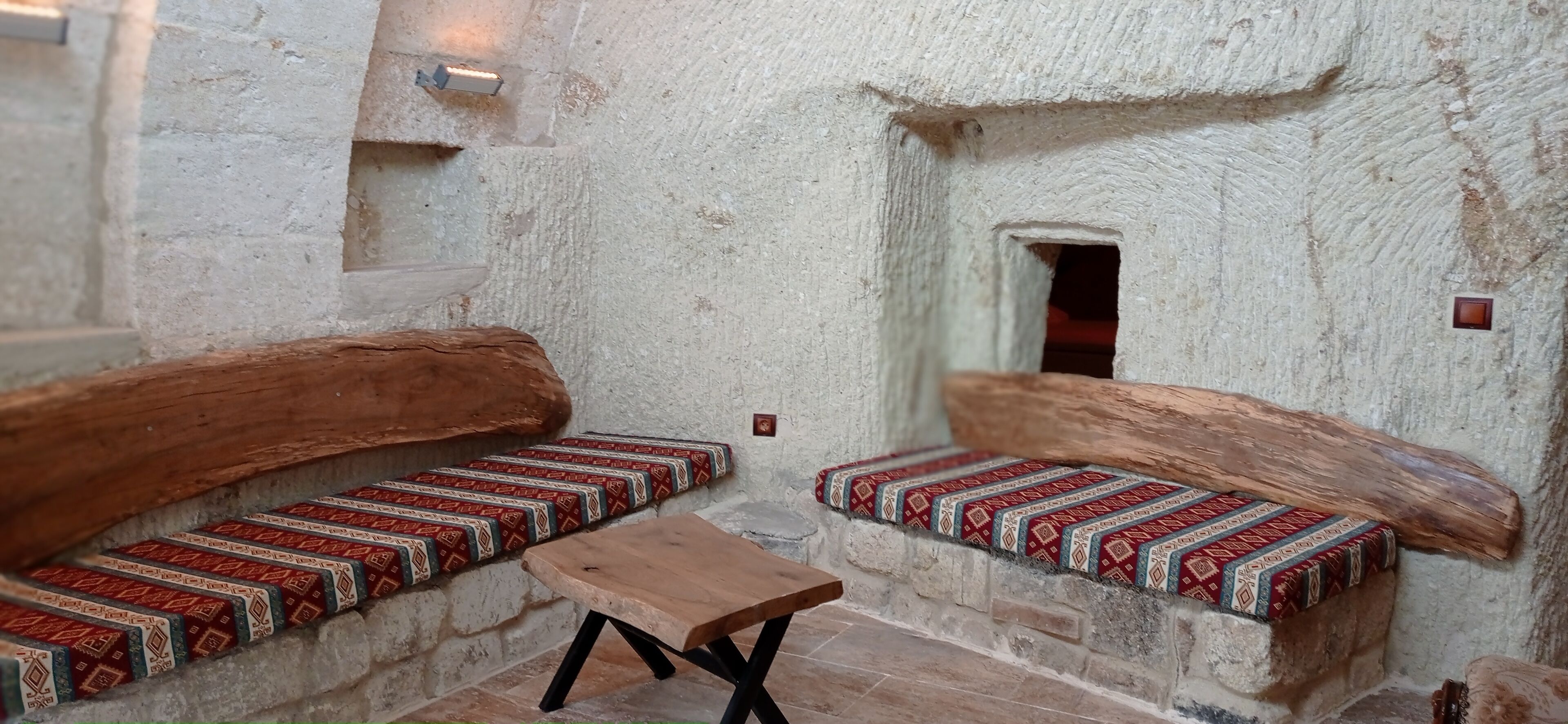A rustic interior with wooden benches and a small table, a stone wall, and a small window.
