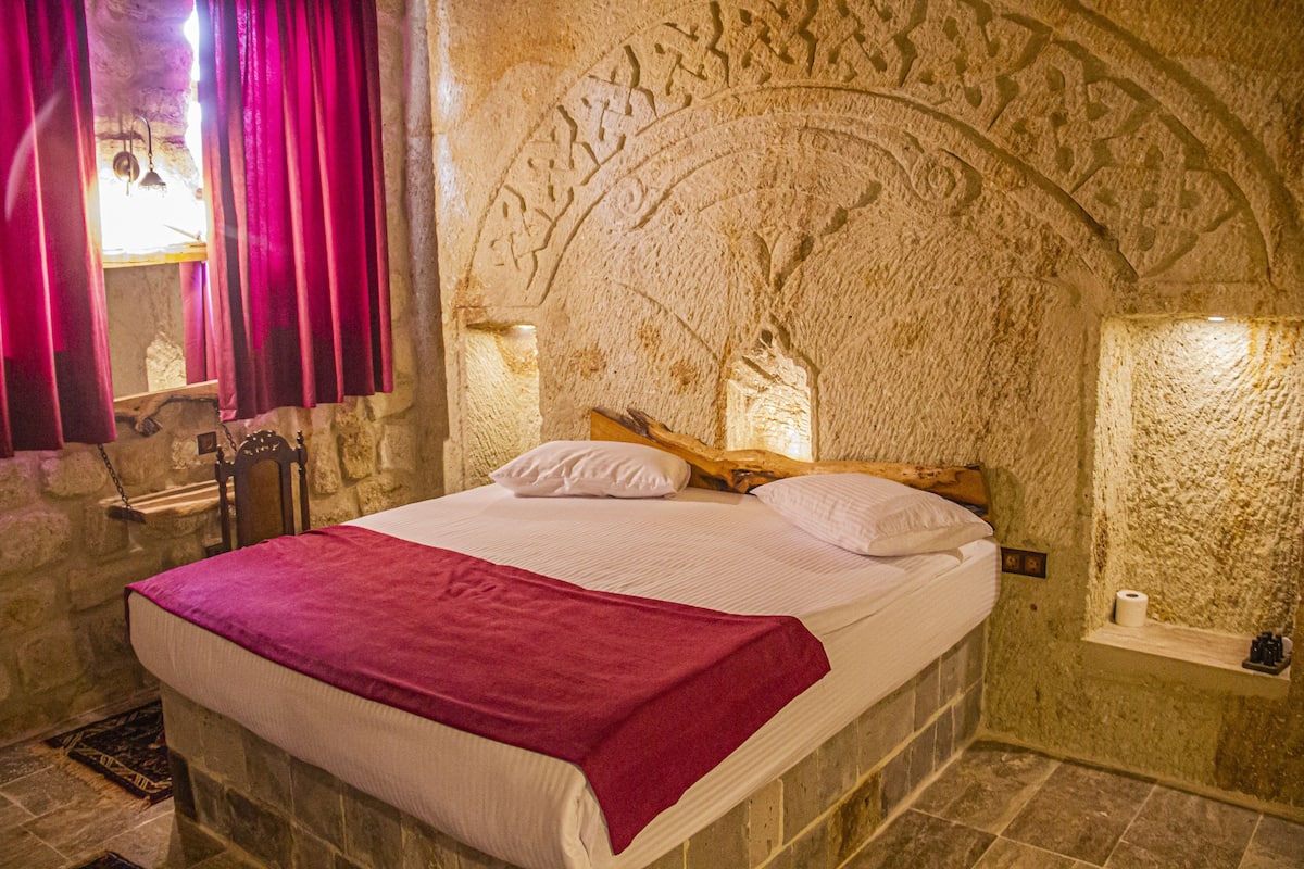 Dormitorio con paredes de piedra, una cama, cabecero de madera y cortina roja.
