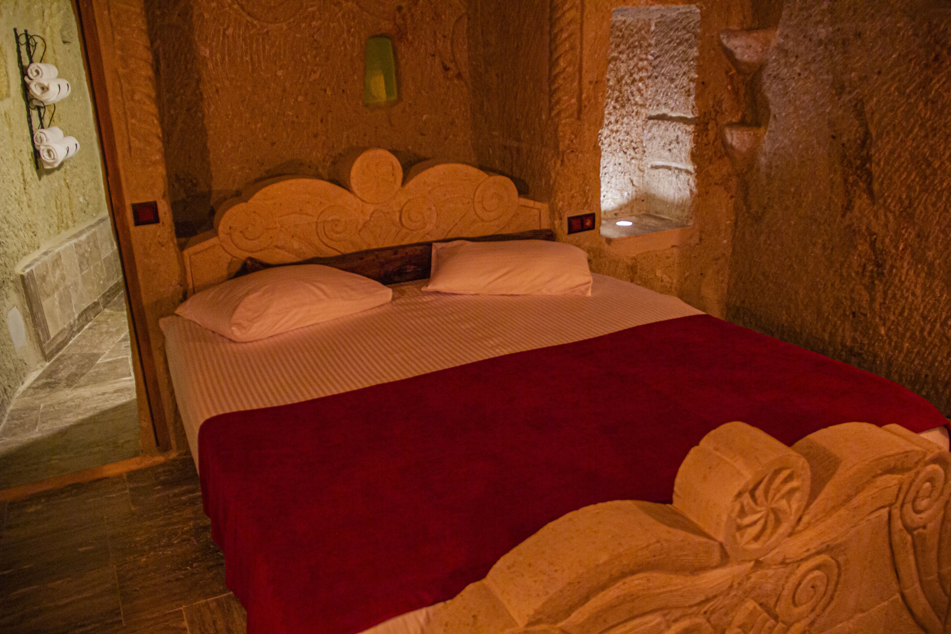 A stone-walled room with a bed, a red blanket, and a carved headboard.