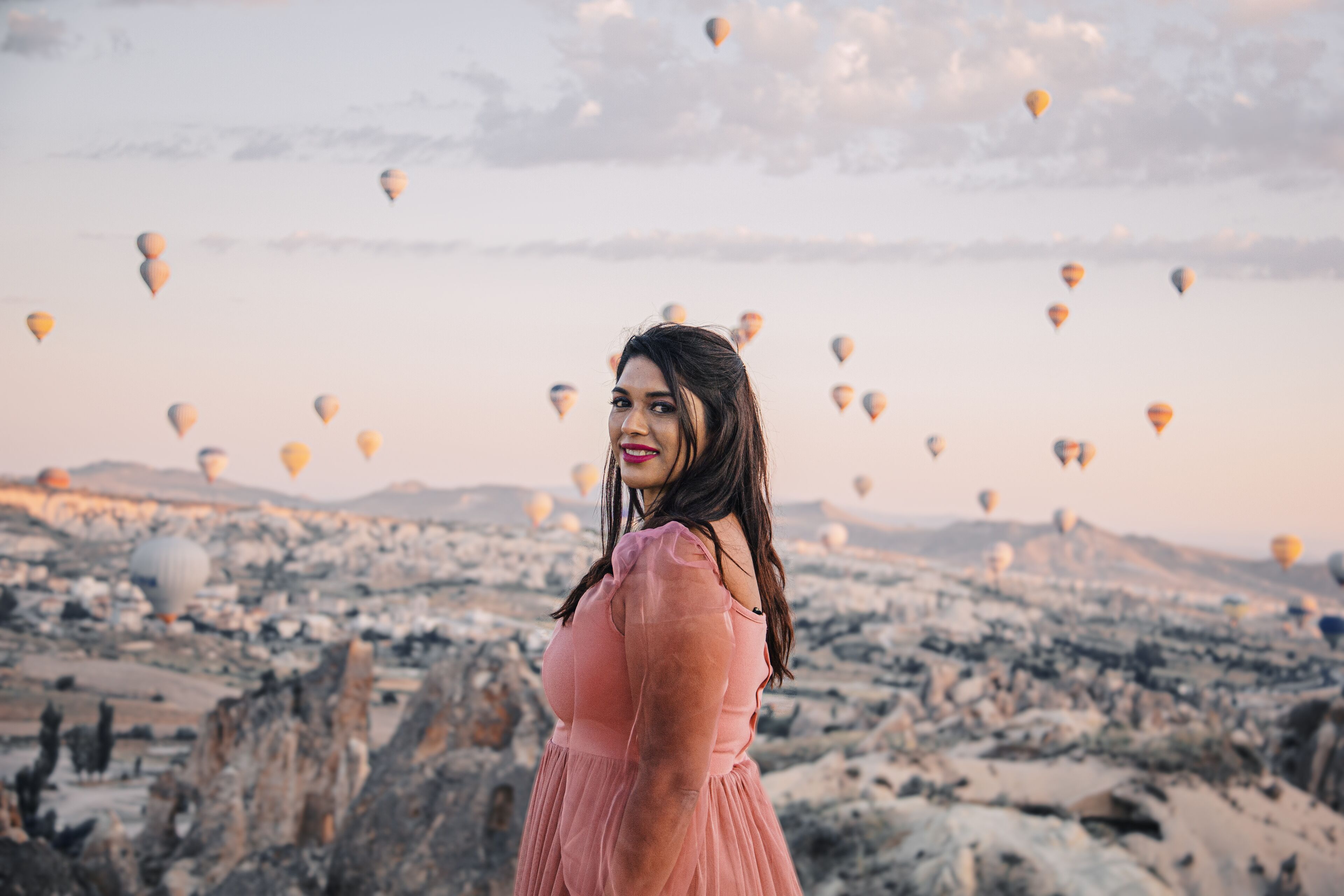 Una persona de pie en un paisaje rocoso con numerosos globos aerostáticos al fondo.
