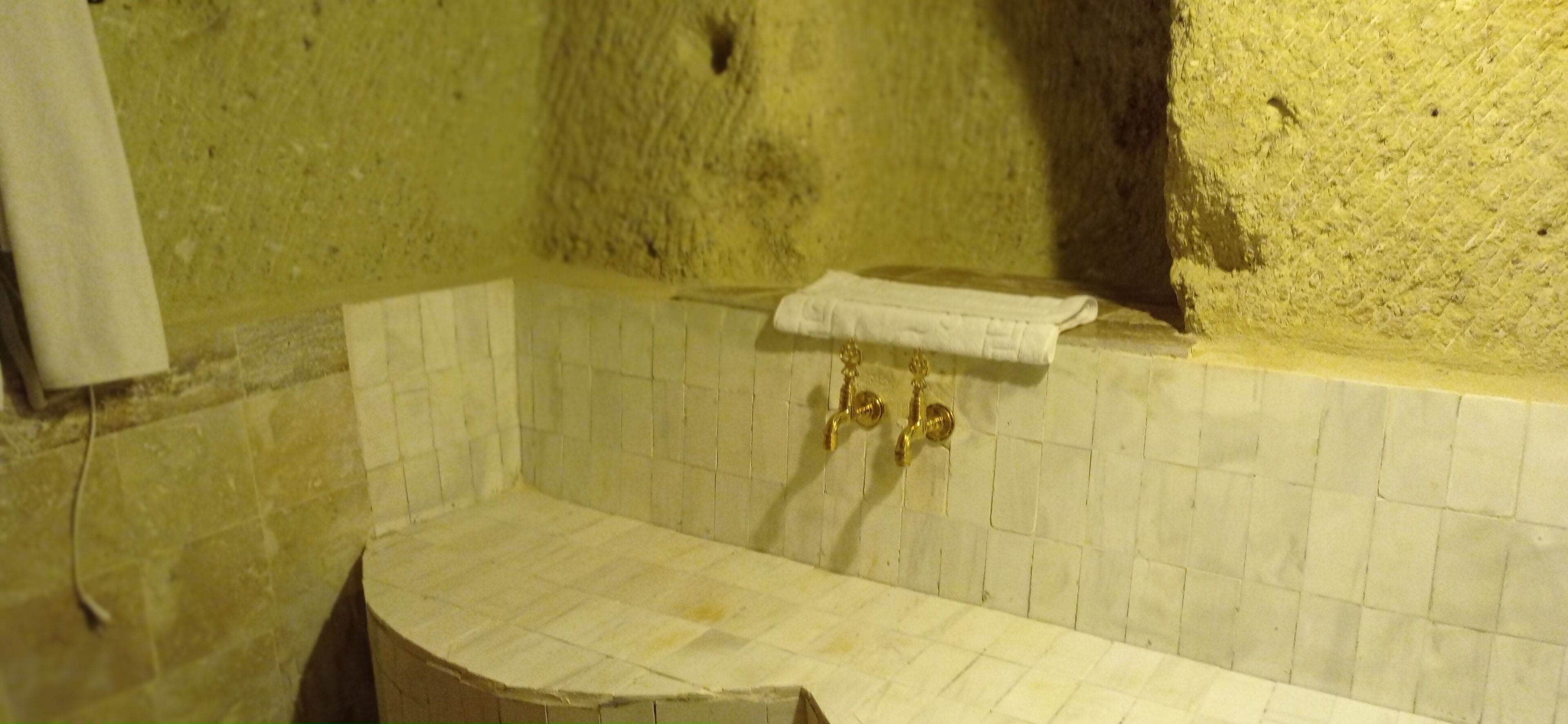Un baño con pared de azulejos blancos, una toalla colgada en un gancho y un grifo dorado.