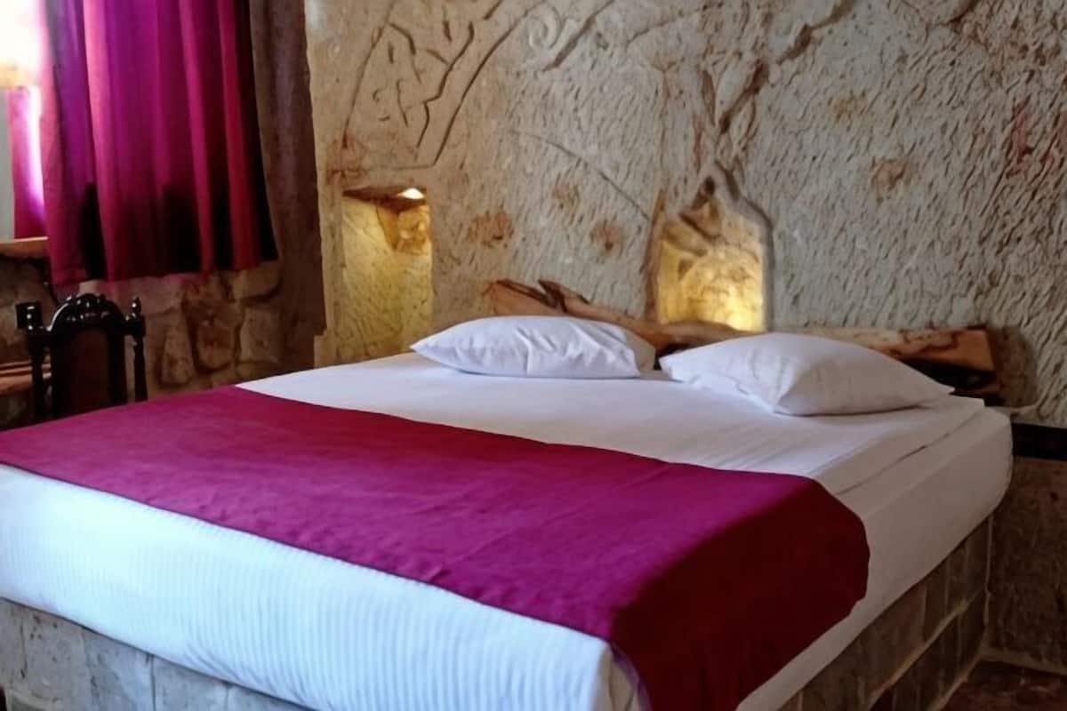 Una cama con ropa de cama blanca y roja, una pared de piedra y una silla de madera.