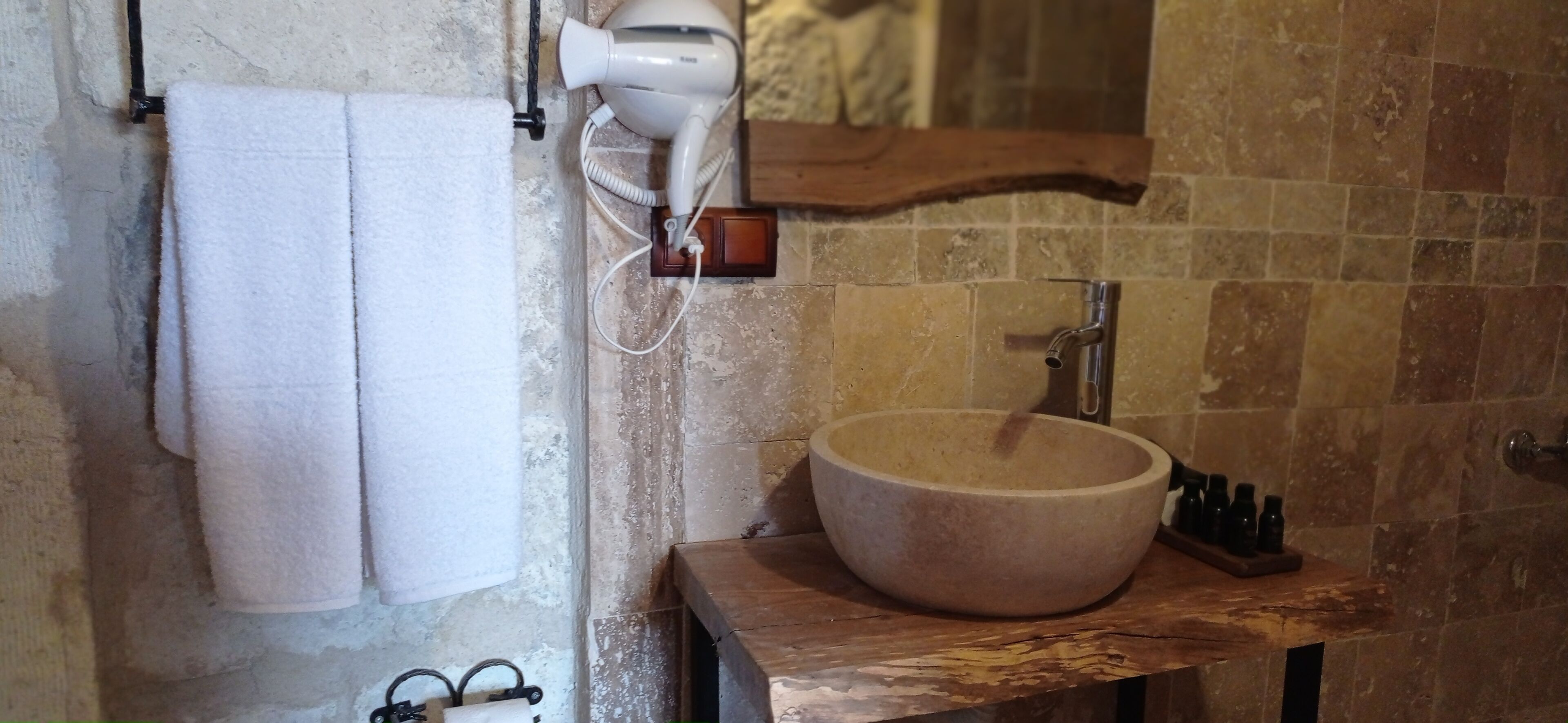 A bathroom with a stone sink, a towel rack with two towels, a hairdryer, and a wooden shelf with bottles.
