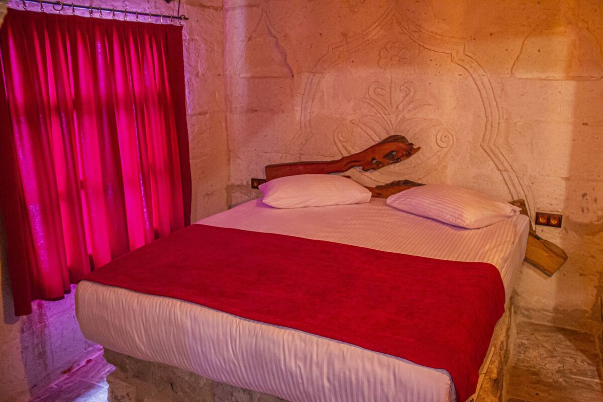 Una cama con una manta roja y almohadas blancas en una habitación con paredes de piedra y un cabecero de madera.