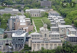 An aerial view of a campus with multiple buildings, including a large central building with a lawn in front.