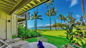 A covered outdoor seating area with a table and chairs, overlooking a well-maintained lawn and palm trees. - Lae Nani 313, Great View Ground Floor (Kapaʻa)