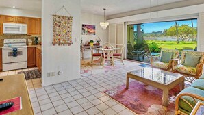 A modern kitchen with white appliances, a dining area with a table and chairs, and a living room with a sofa and a view of a green landscape. - Lae Nani 313, Great View Ground Floor (Kapaʻa)