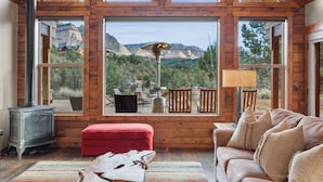 A spacious living room with a large window offering a mountain view, a wooden ceiling, and a red fan. - Skyfall Cabin | Private Hot tub | Zion NP (Orderville)