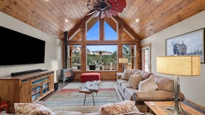 A spacious living room with a large TV, wooden furniture, a red ottoman, and a view of the mountains through large windows. - Skyfall Cabin | Private Hot tub | Zion NP (Orderville)