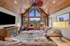 Amplia sala de estar con un televisor grande, muebles de madera, un otomano rojo y vistas a las montañas a través de ventanas grandes.