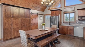 A kitchen with wooden cabinets, a dining table with benches, a chandelier, and a view of the outdoors. - Skyfall Cabin | Private Hot tub | Zion NP (Orderville)