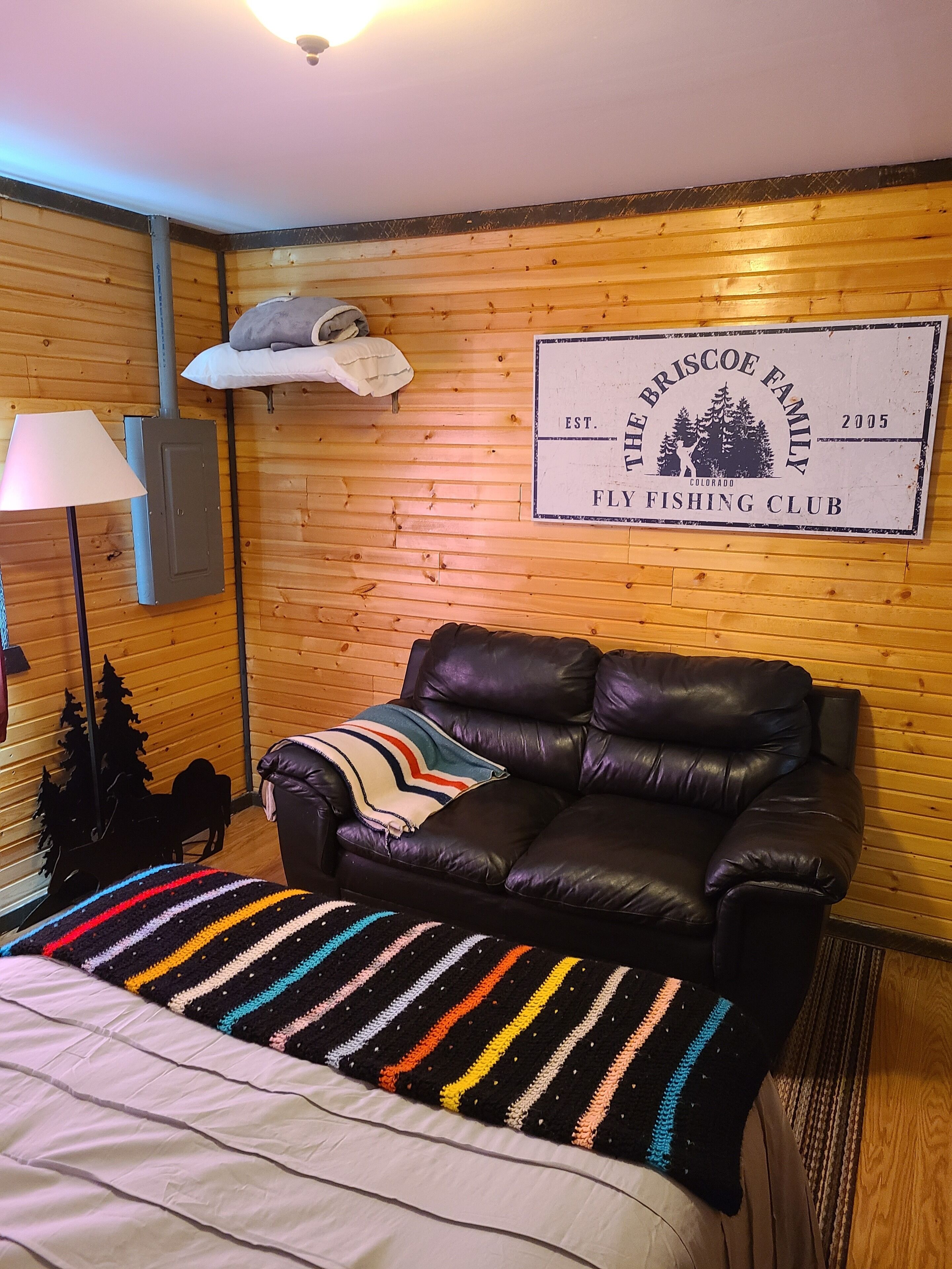 A room with a black leather sofa, a colorful striped blanket, a lamp, and a sign on the wall.