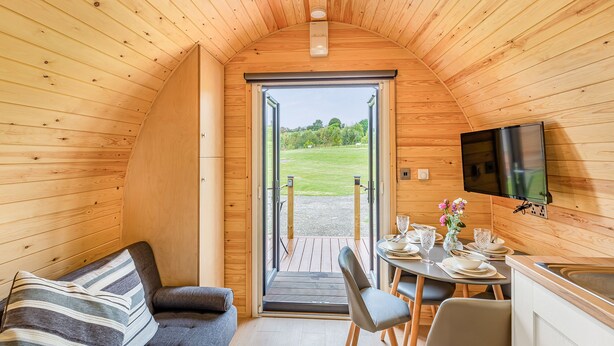 Interior acogedor de una cabaña con techo de madera, un sofá, una mesa de comedor con sillas y un televisor montado en la pared.