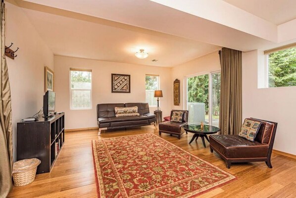 A spacious living room with a large wooden floor, a dark brown sofa, a red patterned rug, and a glass coffee table.