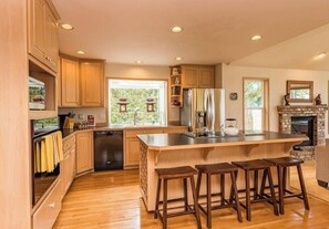 A modern kitchen with wooden cabinets, a large island with stools, a built-in oven, and a stone fireplace.