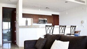 A modern living room with a dark brown sofa, a white pillow, and a kitchen area with wooden cabinets and stainless steel appliances. - BEACHFRONT 2 BEDROOM CONDO IN LAS GAVIAS RESORT - Sleeps 6 (Mazatlán)
