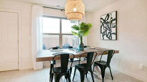 Ein Essbereich mit einem Holztisch, vier schwarzen Stühlen, einer hängenden Rattan-Leuchte, einem Blumentopf und einem gerahmten abstrakten Gemälde an der Wand.