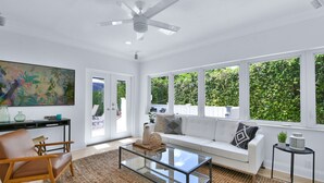 A spacious living room with a glass coffee table, a white sofa with cushions, a large painting on the wall, and a view of the outdoors through large windows.