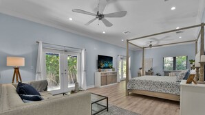 A spacious bedroom with a large bed, a ceiling fan, a TV, and a balcony with a view of greenery.