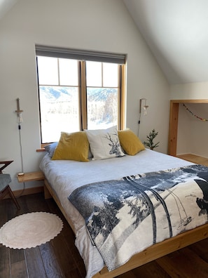 A bedroom with a bed, a window with a view of mountains, a bedside table with a lamp, and a small plant.