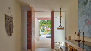 A modern hallway with a wooden door, a macrame wall hanging, a hanging light fixture, and a view of the outside. - Casa Kux  -  Yucatan Home Rentals (Chicxulub Puerto)