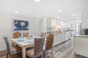 A dining area with a table set for four, a kitchen with white cabinets, and a wall-mounted artwork.
