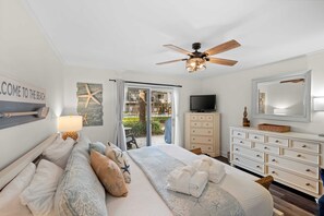 A bedroom with a bed, a ceiling fan, a TV, a dresser, and a view of the beach.