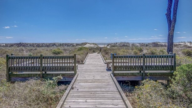 Une promenade en bois mène à deux bancs également en bois, le tout étant entouré d’une végétation naturelle et surplombé par un ciel bleu clair.