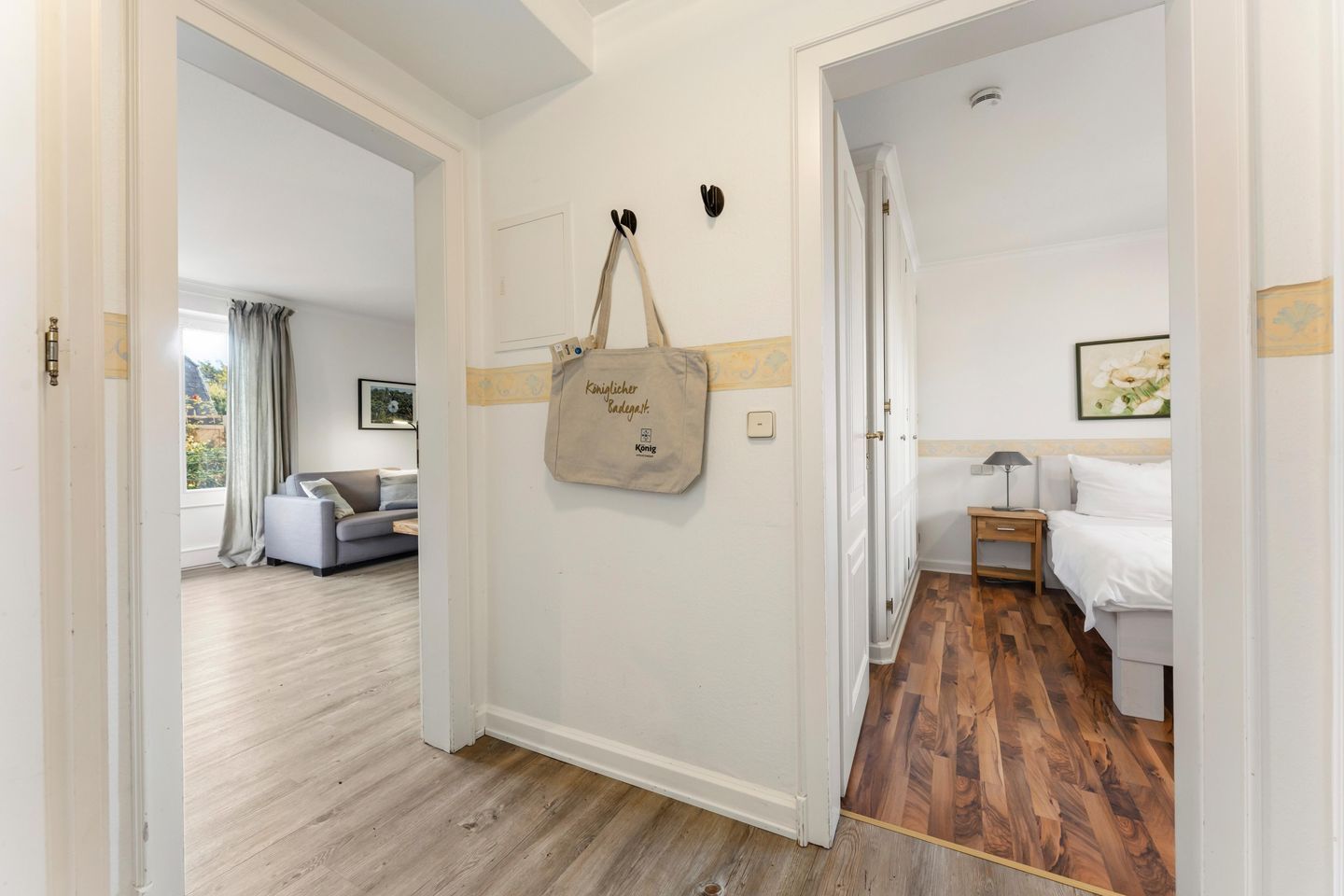 A hotel room with a wooden floor, a bed with white bedding, a nightstand, a lamp, and a wall-mounted bag.