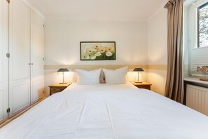 A hotel room with a large bed, two bedside tables with lamps, a painting on the wall, and a window with curtains. - Senwai-Hüs, Apartment 1, Braderup - Senwai-Hüs (Sylt)