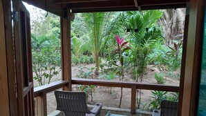 Uma área de estar coberta ao ar livre com duas cadeiras de vime e uma mesa pequena, com vista para um jardim com diversas plantas e árvores.