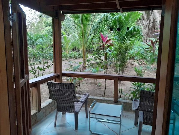 A covered outdoor seating area with two wicker chairs and a small table, overlooking a garden with various plants and trees.
