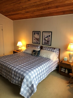 A bedroom with a checkered bedspread, two pillows, a nightstand with a lamp, and two framed posters on the wall.