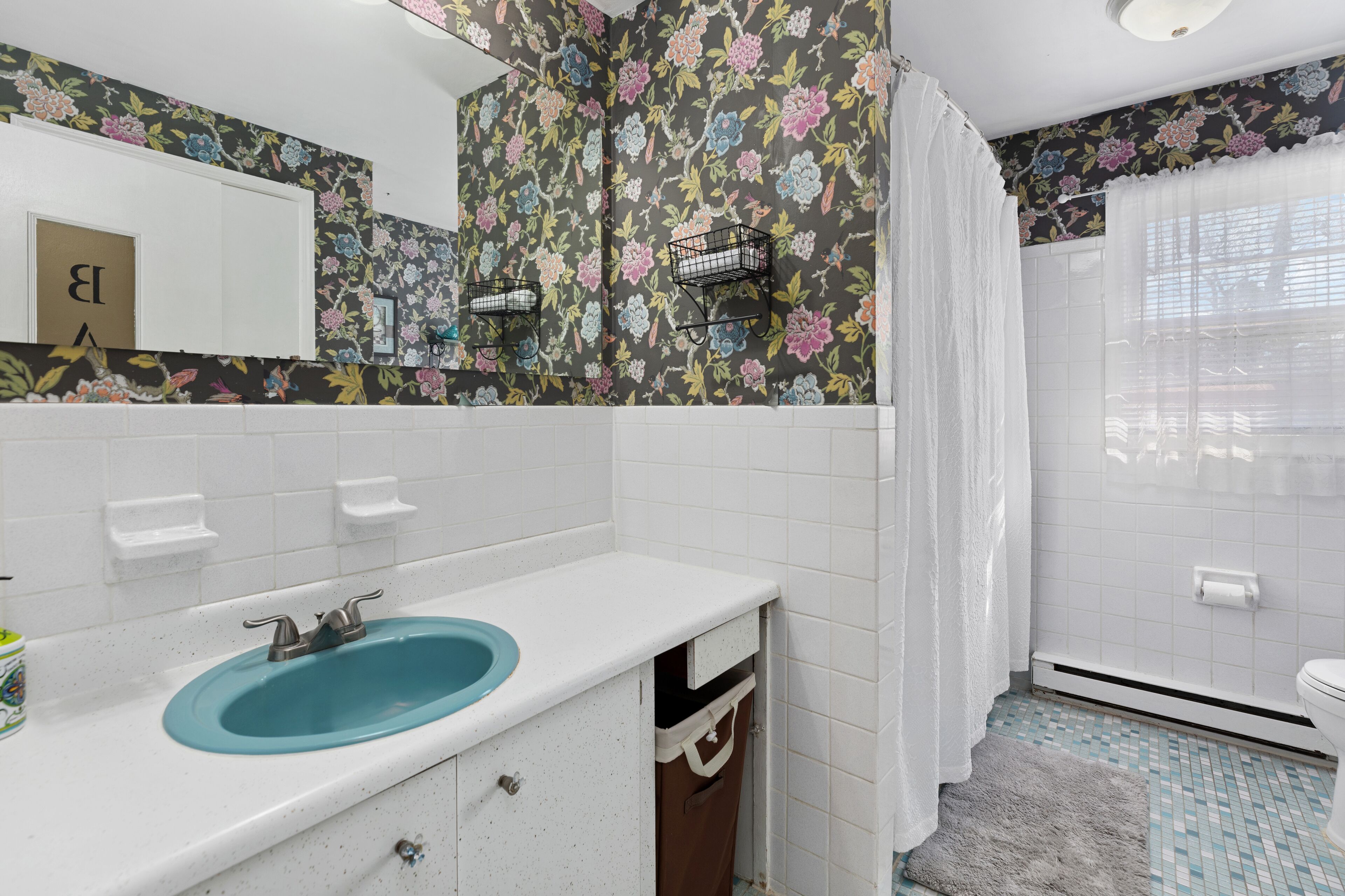 Une salle de bain avec un lavabo bleu, des carreaux blancs, un papier peint à motifs floraux et un espace douche.