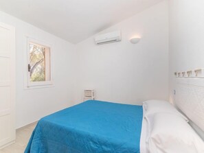 A bedroom with a bed, white walls, an air conditioning unit, a small white shelf, and a window with a view of trees.