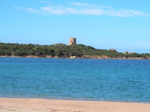 Eine Küstenlandschaft mit einem Turm auf einem Hügel, klarem, blauem Wasser und einem Sandstrand.