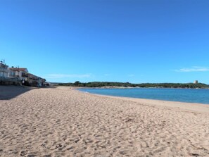 A sandy beach with a clear blue sky, a calm sea, and a building with multiple balconies on the left.