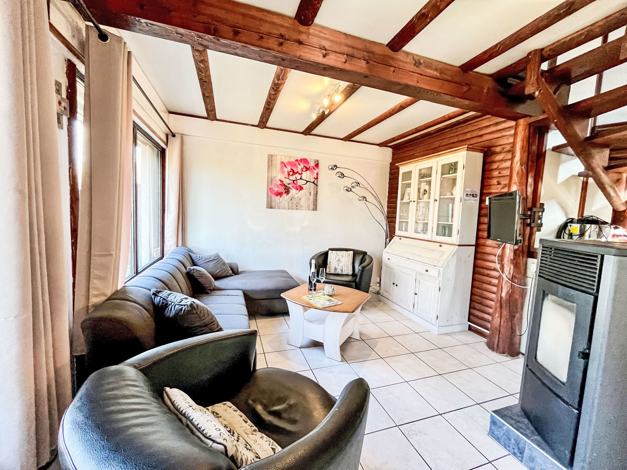 A cozy living room with a sofa, armchair, and a small table. There is a cabinet with glass doors and a television. The room has wooden beams on the ceiling and a tiled floor.
