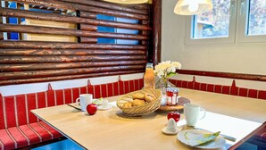 Ruokailutila, jossa on puinen penkki, leipäkori pöydällä, lasi mehua ja maljakko kukkineen.