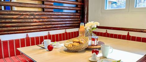 A dining area with a wooden bench, a table with a basket of bread, a glass of juice, and a vase with flowers.
