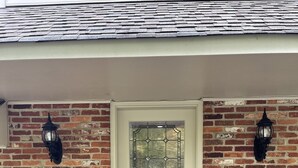A brick house with a white front door, two black lantern-style wall lights, and a pair of metal chairs on the left side. - Roomy 2 Bedroom Apt. w/en Suite BA, Private Entry & Parking, Tennis Court (Bethesda)