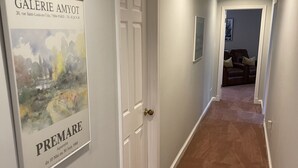 A hallway with a carpeted floor, white walls, and a door. There is a framed picture on the wall and a chair in the background. - Roomy 2 Bedroom Apt. w/en Suite BA, Private Entry & Parking, Tennis Court (Bethesda)