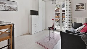 Uma sala de estar moderna com um sofá, uma mesa de jantar com cadeiras, uma pequena mesa lateral e um armário branco. Há uma janela com vista para edifícios e uma porta que dá para uma área externa.