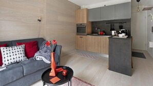 Um apartamento moderno com sofá cinzento, uma pequena mesa lateral e uma área de cozinha com armários cinzas e bancada preta.
