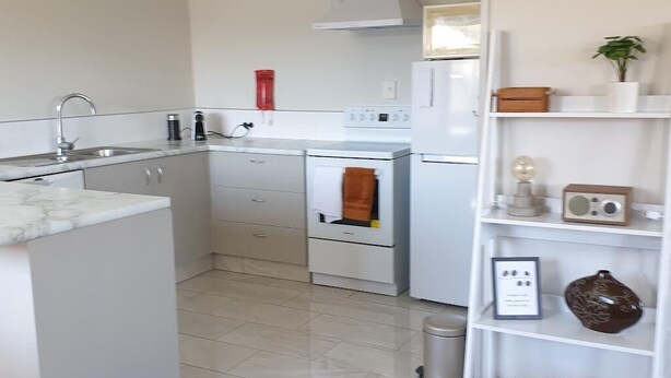 A modern kitchen with white cabinetry, a marble countertop, a built-in oven, and a white shelving unit with decorative items. - Kiwiana Gem to enjoy in Reefton (Reefton)