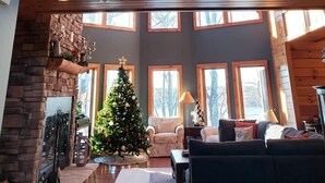 A spacious living area with a high ceiling, wooden beams, a stone fireplace, a Christmas tree, and large windows.