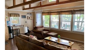 Una sala de estar acogedora con un sofá, sillones y una mesita de centro. Se ve una ventana con persianas, un dispensador de agua y una pequeña mesa con una planta.