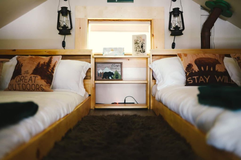 Een gezellige kamer met twee houten stapelbedden, elk voorzien van een kussen met tekst over het buitenleven. In het midden staat een plankje met een ingelijste foto en een kleine plant.